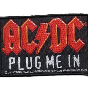 May include: Black embroidered patch featuring the band name "AC/DC" in red and yellow lettering, along with the phrase "PLUG ME IN" in white lettering. The patch includes the copyright information "2019 Leid Seplein Presse B.V. under license to Perryscope Productions/Epic Rights."