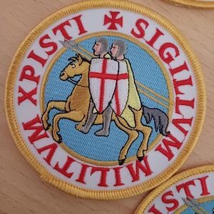 Puede incluir: Un parche redondo bordado con fondo blanco y borde dorado. En el centro, dos caballeros a caballo, una cruz roja y el texto "XPISTI SIGILLVM WLITIVM".