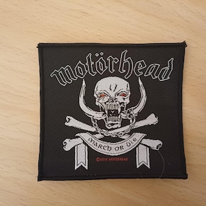 Puede incluir: Un parche bordado en blanco y negro con el nombre de la banda "Motörhead" en una fuente gótica. Una calavera con ojos rojos y una cadena alrededor del cuello está rodeada de huesos cruzados y una pancarta que dice "March or Die". El texto "©2010 Motörhead" está en la parte inferior de la pancarta.