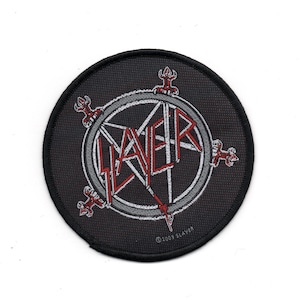 May include: A black and white embroidered patch featuring the Slayer logo. The logo is a circle with a pentagram inside and the word "Slayer" written in red letters. The patch has a black border and the text "©2003 Slayer" at the bottom.
