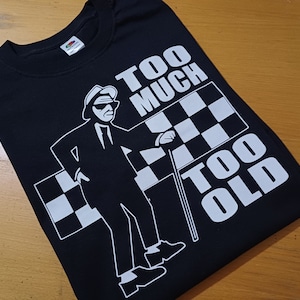 Op de afbeelding: Zwart T-shirt met een witte afbeelding van een man in pak met een stok en een geruite vlag. De tekst "Too Much Too Old" staat op het shirt gedrukt.