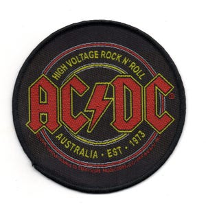 Puede incluir: Un parche circular negro y amarillo con el texto "HIGH VOLTAGE ROCK N' ROLL" y "AC/DC" en letras rojas y amarillas. El texto "AUSTRALIA EST. 1973" también está incluido en letras amarillas.