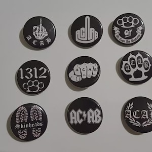 Op de afbeelding: Negen zwart-witte speldjes met diverse ontwerpen. De speldjes tonen afbeeldingen van een middelvinger, boksbeugels en tekst, waaronder "ACAB", "1312" en "Skinheads".