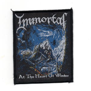 Könnte beinhalten: Schwarzer und weißer gestickter Patch mit dem Bandnamen "Immortal" in einer stilisierten Schriftart. Der Patch zeigt eine wolfähnliche Kreatur in einer schneebedeckten Landschaft mit dem Text "At The Heart Of Winter".