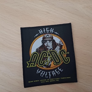 Puede incluir: Un parche de tela negro con un círculo amarillo que presenta una imagen de dibujos animados de una persona con una gorra de béisbol y sosteniendo una guitarra. El texto "HIGH VOLTAGE" y "AC/DC" están en letras amarillas.