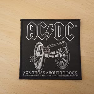 Puede incluir: Parche bordado en blanco y negro con el nombre de la banda "AC/DC" y el texto "For Those About To Rock" con un gráfico de cañón. ©2015 Under License to Perryscope Productions LLC/Epic Rights Inc.