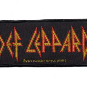 以下が含まれることがあります： バンド名「Def Leppard」が黄色と赤の文字で書かれた黒い織りパッチ。テキストは鋭角とギザギザのフォントでスタイリッシュにデザインされています。著作権情報「©2020 Bludgeon Riffola Limited」は、バンド名の下に白で印刷されています。