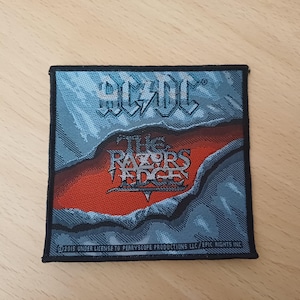 Könnte beinhalten: Quadratischer Aufnäher mit dem AC/DC-Logo oben und "The Razors Edge" in der Mitte. Der Hintergrund ist blau mit einem zerrissenen Papiereffekt, der einen roten Bereich freilegt. Schwarzer Rand.