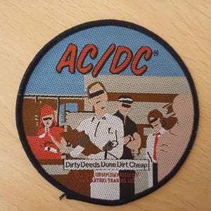 May include: A circular embroidered patch featuring the band AC/DC with the text "Dirty Deeds Done Dirt Cheap". The patch features a cartoon image of five people, including a person in a red hat and a person in a white shirt.