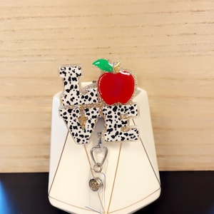 May include: A badge reel with the word "LOVE" in a dalmatian print design, with a red apple and green leaf accent. The apple is embellished with glitter. The reel has a clear plastic holder and a silver-tone clip.