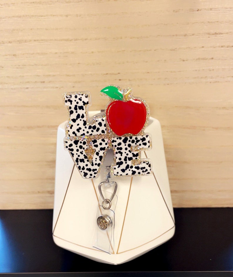 May include: A badge reel with the word "LOVE" in a dalmatian print design, with a red apple and green leaf accent. The apple is embellished with glitter. The reel has a clear plastic holder and a silver-tone clip.