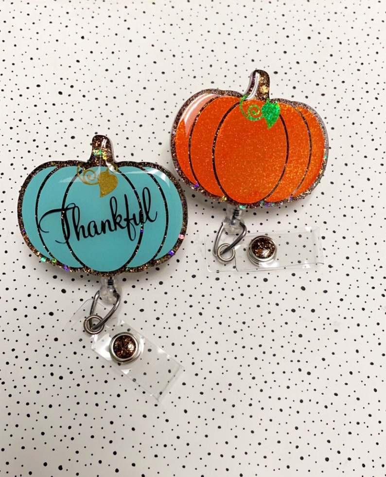 May include: Two pumpkin-shaped badge reels. One is teal with the word "Thankful" in script. The other is orange. Both have glitter accents, a clear plastic reel, and a metal clip. The background is white with black polka dots.