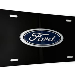 May include: A black license plate featuring the Ford logo. The logo is an oval with a silver border, a blue center, and the word "Ford" in white script. The plate has four mounting holes.