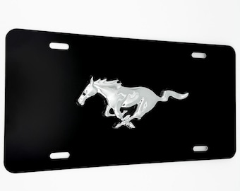 May include: Black license plate with a chrome-colored running horse emblem. The plate has four oblong holes for mounting. The horse is a stylized representation of a Mustang.