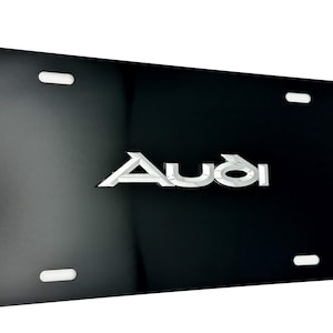 May include: Black license plate with the Audi logo in silver. The plate has four white mounting slots and a glossy finish. The Audi logo is centered and has a reflective appearance.