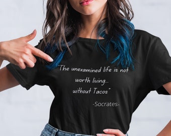May include: A black t-shirt with a white quote that reads "The unexamined life is not worth living... without Tacos" -Socrates-. The person wearing the shirt is pointing to the quote.