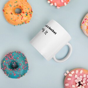 May include: White ceramic mug with black text that reads "in a bucket #*k it". The mug is surrounded by colorful donuts with sprinkles.