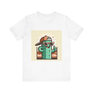 May include: White t-shirt featuring a cartoon cactus wearing sunglasses and a backwards baseball cap. The cactus is giving a thumbs up. The design includes a desert landscape and a setting sun.