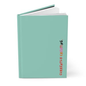 May include: A teal-coloured hardcover journal with the text "CURRENTLY CREATING" in colourful lettering on the spine. The journal is slightly open, revealing white pages. The cover is a solid, light teal colour.
