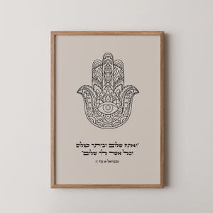 May include: Framed art print featuring a black Hamsa hand design with an eye in the center, set against a beige background. The print includes Hebrew text below the hand, all within a light brown wooden frame.