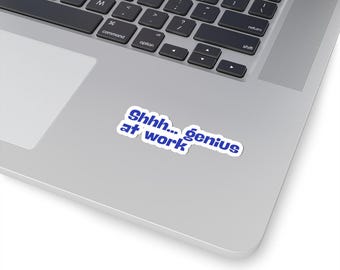 May include: A white sticker with blue text that reads "Shhh... genius at work." The sticker is on a silver laptop, near the keyboard. The laptop is open, and the keyboard is visible.