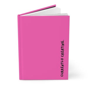 May include: A bright pink journal with the words "CURRENTLY CREATING" printed vertically in black on the right side. The journal is open, revealing white pages.