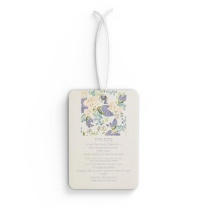 Op de afbeelding: Een rechthoekige luchtverfrisser met een bloemenontwerp met witte en paarse bloemen en groene bladeren. De luchtverfrisser heeft een wit touwtje om op te hangen en tekst in het Hebreeuws.