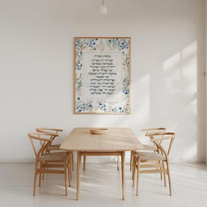 Könnte beinhalten: Ein gerahmter Druck mit einem floralen Rahmen und hebräischem Text. Der Text ist ein Gebet in Hebräisch. Der Druck hängt an einer weißen Wand über einem Holztisch mit zwei Stühlen.