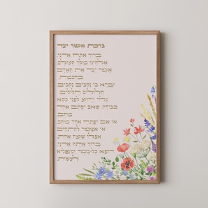 May include: A framed print featuring Hebrew text in gold lettering on a light pink background. The artwork includes a watercolor floral arrangement in the lower right corner, with red poppies, bluebells, and other wildflowers. The frame is a warm, natural wood tone.