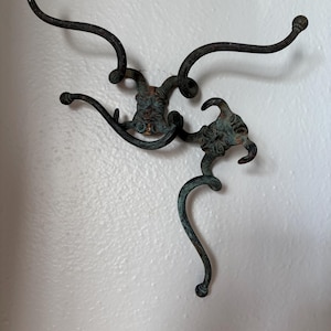 Puede incluir: Dos perchas de pared antiguas de bronce con un diseño decorativo. Cada gancho presenta un elemento central ornamentado con brazos curvos que terminan en pequeñas puntas esféricas. Los ganchos están montados en una pared blanca.