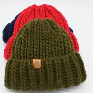 Könnte beinhalten: Drei grob gestrickte Mützen in Olivgrün, Rot und Marineblau sind auf einer weißen Oberfläche gestapelt. Die olivgrüne Mütze befindet sich im Vordergrund, mit einem kleinen braunen Etikett. Die Mützen sind aus dickem, strukturiertem Garn.
