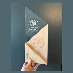 May include: A clear acrylic and wood trophy with the text "CANADIAN SWING CHAMPIONSHIPS" and "Cabaret 2024". The trophy is engraved with a laurel wreath and "1st place".