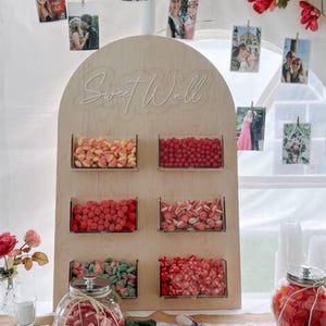 Può includere: Un espositore in legno "Sweet Wall" con contenitori trasparenti pieni di caramelle colorate. L'espositore è decorato con foto, fiori e un cuore di legno con la scritta "Love". Vasi di vetro con caramelle e tovaglioli sono sul tavolo.
