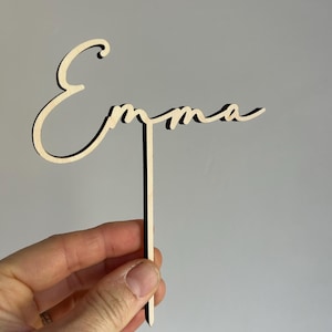Peut inclure: Un topper de gâteau en bois avec le nom "Emma" écrit en cursive. Le topper est fixé à un bâtonnet en bois fin.