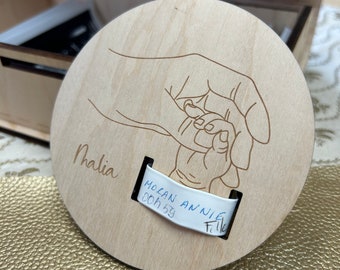 Può includere: Un cerchio di legno con un design inciso a laser di due mani che si tengono. Il cerchio ha il nome "Malia" scritto su di esso e un'etichetta bianca con il testo "Molan Annie 00h 59 File" scritto su di essa.