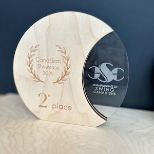 May include: A wooden trophy with a clear acrylic accent. The trophy is round with a crescent-shaped cutout. Text on the wooden portion reads "Canadian Showcase 2025" and "2nd place". The acrylic section has text that reads "Championnats de Swing Canadiens".