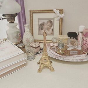 May include: A gold-colored Eiffel Tower figurine stands on a white surface, surrounded by perfume bottles, a framed picture, and decorative items. A vintage lamp and a snow globe are also visible, creating a romantic, feminine aesthetic.