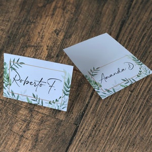May include: Two white place cards with green foliage and gold frames. The text on the cards reads "Roberto F." and "Amanda D."