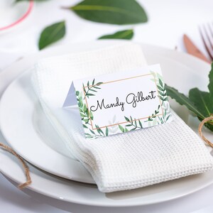 May include: A white place setting with a white napkin, a white plate, and a place card with the name "Mandy Gilbert" printed on it. The place card has a green and gold floral design.