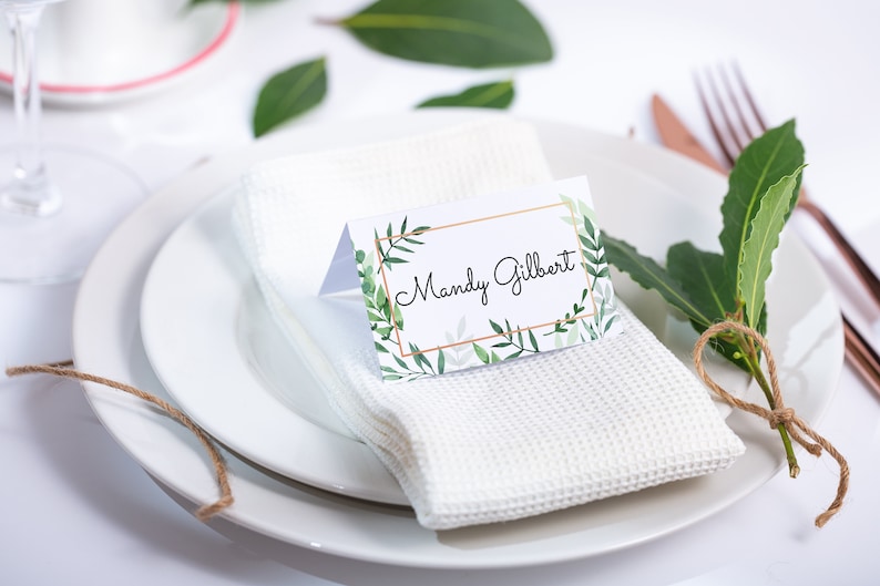 May include: A white place setting with a white napkin, a white plate, and a place card with the name "Mandy Gilbert" printed on it. The place card has a green and gold floral design.