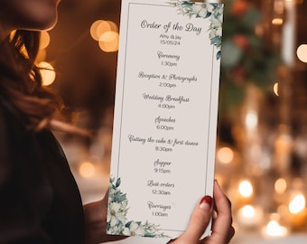 May include: A white wedding order of the day card with green floral accents. The card lists the wedding events with times, including the ceremony, reception, wedding breakfast, speeches, cutting the cake, supper, last orders, and carriages.