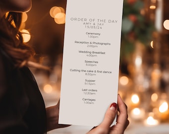 May include: A white card with the text "ORDER OF THE DAY" at the top, followed by "AMY & JAY" and "15/05/24". The card lists the order of events for a wedding day, including the ceremony, reception, wedding breakfast, speeches, cutting the cake and first dance, supper, last orders, and carriages.
