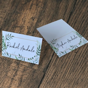 May include: Two white place cards with gold frames and green watercolor leaves. The text on the cards reads "Rachel Andale" and "Stephen Andale".