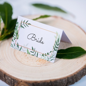May include: White place card with a gold frame and green foliage design. The text "Bride" is written in black cursive inside the frame.