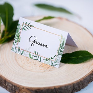 May include: White wedding place card with a gold and green leafy design and the word "Groom" in black cursive lettering.