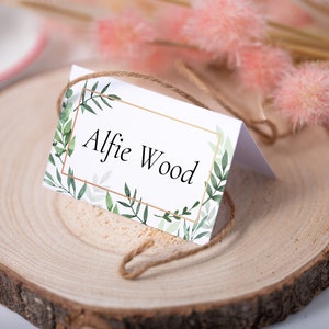 May include: A white place card with a gold frame and green foliage design. The text "Alfie Wood" is printed in black inside the frame.
