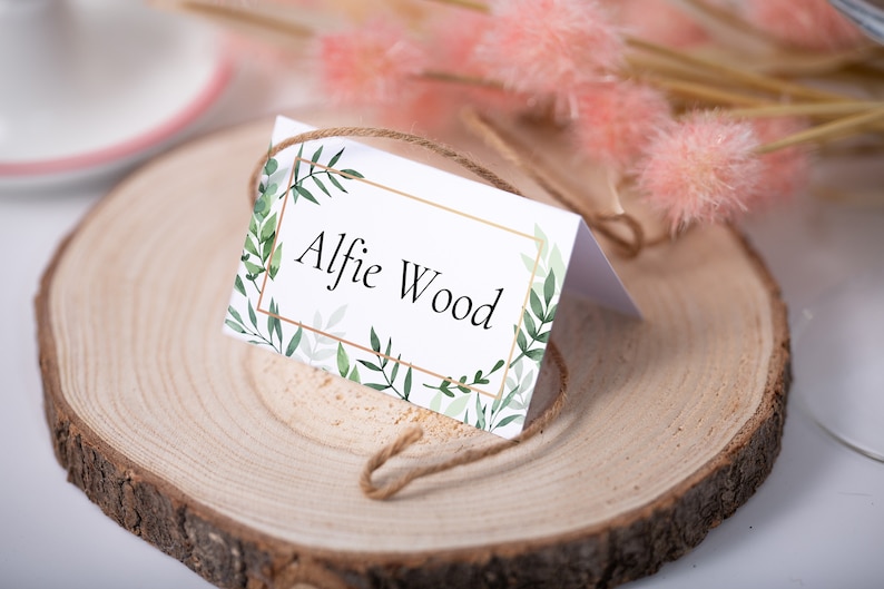 May include: A white place card with a gold frame and green foliage design. The text "Alfie Wood" is printed in black inside the frame.