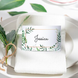 May include: A white place card with a gold frame and green foliage design. The name "Jessica" is written in black cursive inside the frame.