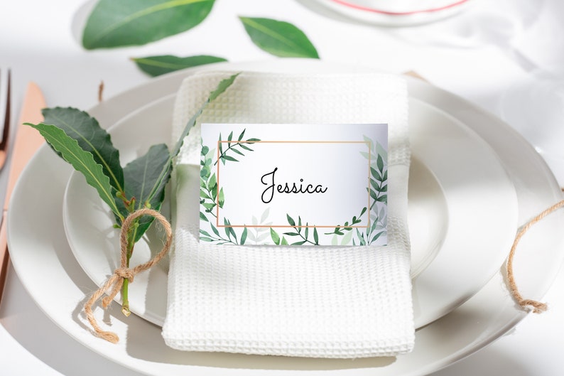 May include: A white place card with a gold frame and green foliage design. The name "Jessica" is written in black cursive inside the frame.