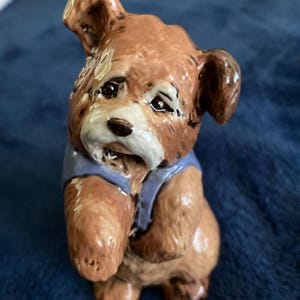May include: A ceramic figurine of a brown puppy with a blue vest. The puppy is in a seated position with its paws together, and it has a sad expression on its face. The figurine has a glossy finish.